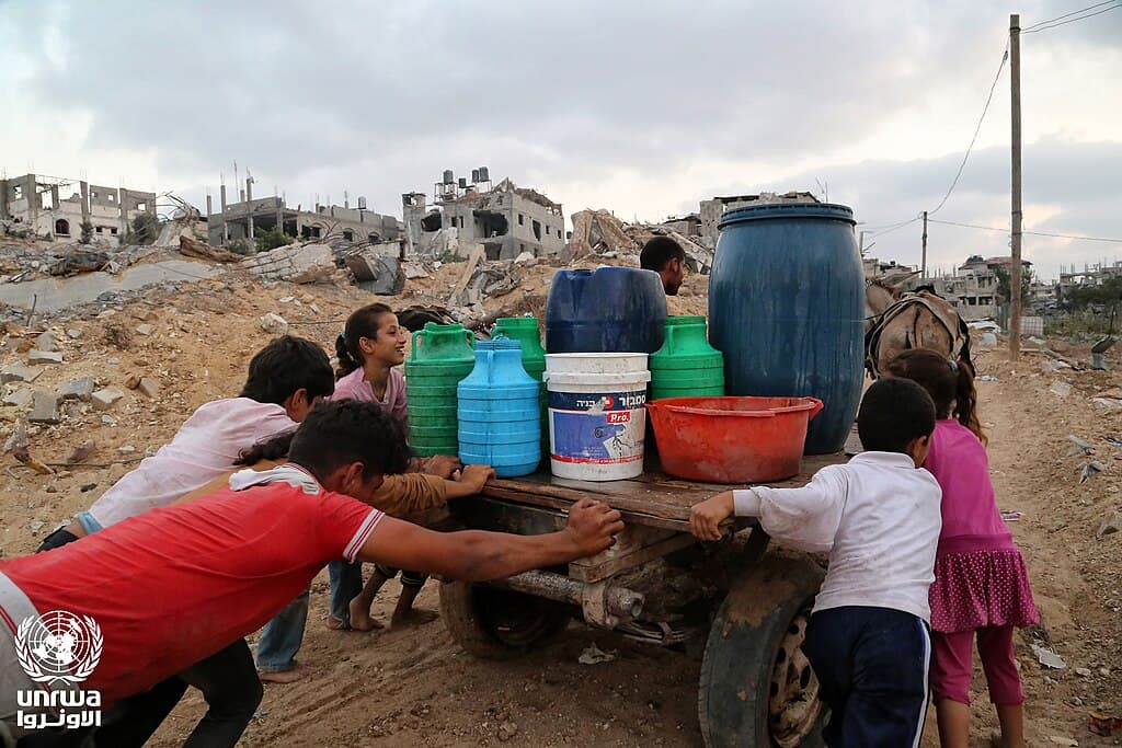 Niños empujan un carro cargado de recipientes de agua entre edificios destruidos en Gaza